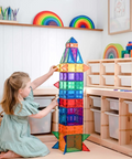 A young girl in a light green dress kneels on the floor of a bright children's room, building a tall translucent rainbow magnetic block tower with a small rocket-shaped top. The room features light wood shelves, white paneling, and colorful toys.