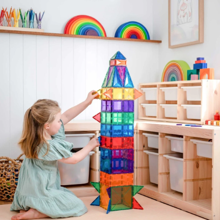 A young girl in a light green dress kneels on the floor of a bright children's room, building a tall translucent rainbow magnetic block tower with a small rocket-shaped top. The room features light wood shelves, white paneling, and colorful toys.