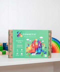 Connetix Rainbow Magnetic Tiles box on a white shelf, with colorful toy blocks and a rainbow arch in the blurred background.