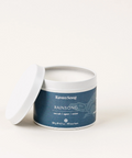 A Ravensong branded candle with the name Rainsong, featuring a white candle in a container with a label that includes amber, jasmine, and tonka scents.