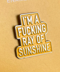 Enamel pin with text 'I'm a fucking ray of sunshine' on a beige background