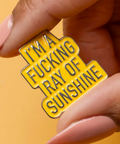 Yellow enamel pin with black text held by a hand against a yellow background