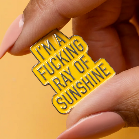 Yellow enamel pin with black text held by a hand against a yellow background