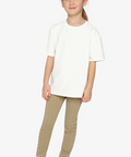 A young girl modeling a white short-sleeve crewneck t-shirt and beige ribbed leggings with white sneakers, standing against a light gray studio background.