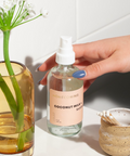 A hand reaching for a clear glass bottle with a white pump labeled 'Honeymoon' filled with a coconut milk and sandalwood scented home mist, next to a vase with flowers and a small container of cotton swabs.