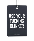 A black double-sided air freshener with the phrase 'USE YOUR FUCKING BLINKER' written in white. It includes a nylon hanging cord.