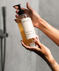 A person's hands holding a pump bottle of body wash with the label 'PICOZ honey tobacco'. The body wash is being squeezed out of the bottle, and there are droplets of water and soap around, indicating it is being used in the shower.