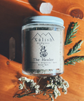 A reusable glass jar labeled 'Xalish medicine The Healer' containing bath salts with a wooden background and decorative plants around it.