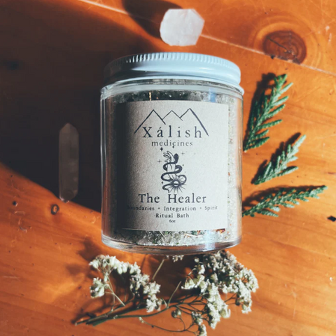 A reusable glass jar labeled 'Xalish medicine The Healer' containing bath salts with a wooden background and decorative plants around it.
