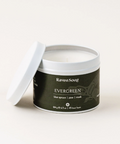 A RavenSong Evergreen candle in a white container with a black label, featuring top notes of blue spruce and musk, displayed against a neutral background.