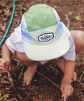 Child wearing a green and white cap with a brand logo, sitting on the ground.