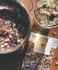 Two metal bowls filled with dried herbs and flowers, a package of Xalishi tea, on a wooden surface.