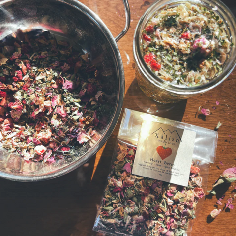 Two metal bowls filled with dried herbs and flowers, a package of Xalishi tea, on a wooden surface.