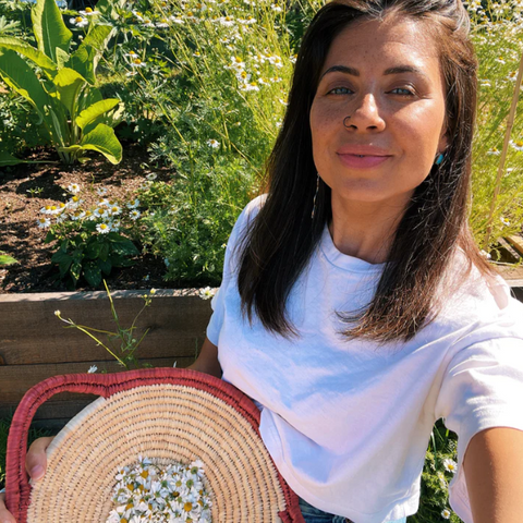 Woman holding a woven bag outdoors with greenery and flowers in the background