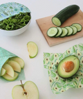 Green reusable food wraps with sliced cucumbers, avocado, and apples on a white background