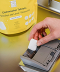 Person inserting a dishwasher tablet into a dispenser with a yellow package labeled 'Dishwasher Tablets' in the background.