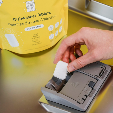 Person inserting a dishwasher tablet into a dispenser with a yellow package labeled 'Dishwasher Tablets' in the background.