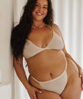 Woman wearing a beige bikini sitting against a white curtain.