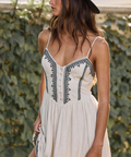 Woman wearing a white dress with black embroidery and a dark hat, standing against a natural background.