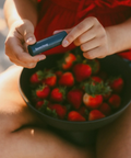 Person holding a bowl of strawberries with a product in their hand, outdoors.