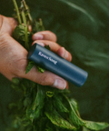 Hand holding a bundle of green leaves with a Raven Song product in the foreground against a green background