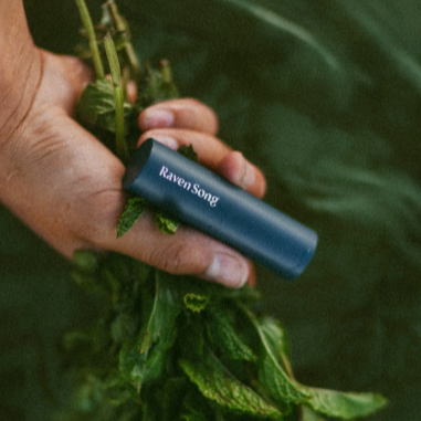 Hand holding a bundle of green leaves with a Raven Song product in the foreground against a green background