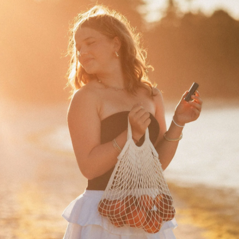 Woman holding a string bag with oranges by a lake at sunset
