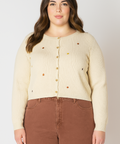 Woman wearing a beige ribbed sweater with colorful buttons and brown pants on a white background