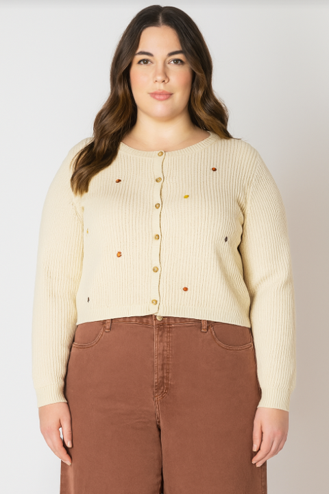 Woman wearing a beige ribbed sweater with colorful buttons and brown pants on a white background