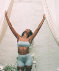 Person in light blue athletic wear standing with arms raised in a room with plants and white walls.
