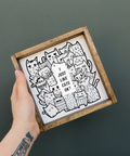 Hand holding a wooden-framed print with black and white cat illustrations on a dark background
