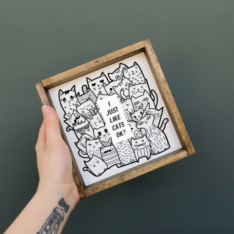 Hand holding a wooden-framed print with black and white cat illustrations on a dark background