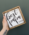 Hand holding a small wooden sign with 'Best Papa Ever' text on a dark background