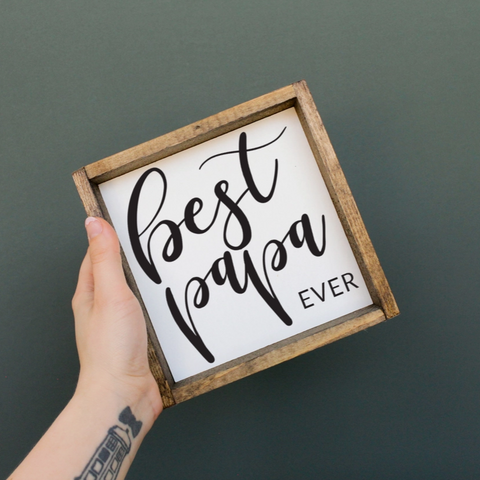 Hand holding a small wooden sign with 'Best Papa Ever' text on a dark background