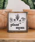 Framed sign with 'plant mom' text and plant illustrations in front of potted plants.