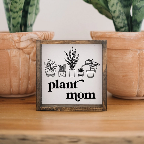 Framed sign with 'plant mom' text and plant illustrations in front of potted plants.