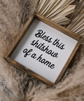 Small wooden sign with text 'Bless this shitshow of a home' on a textured surface.