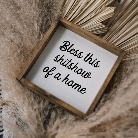 Small wooden sign with text 'Bless this shitshow of a home' on a textured surface.