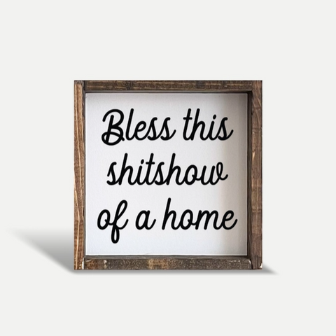 Wooden sign with text 'Bless this shitshow of a home' on a white background
