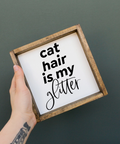 Hand holding a sign with 'cat hair is my glitter' text against a dark background
