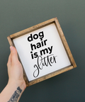 Hand holding a framed sign with 'dog hair is my glitter' text on a dark background