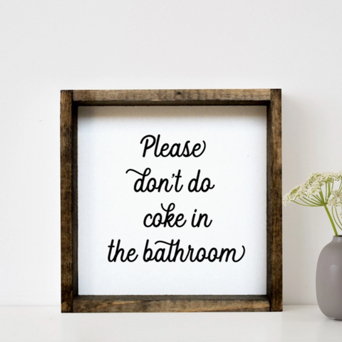 Framed sign with text 'Please don't do coke in the bathroom' on a white wall with a vase and plant.