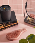 Rose gold hairbrush on a pink surface with books and a candle in the background