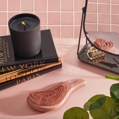 Rose gold hairbrush on a pink surface with books and a candle in the background