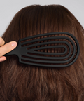 Person using a black hairbrush on their brown hair against a gray background