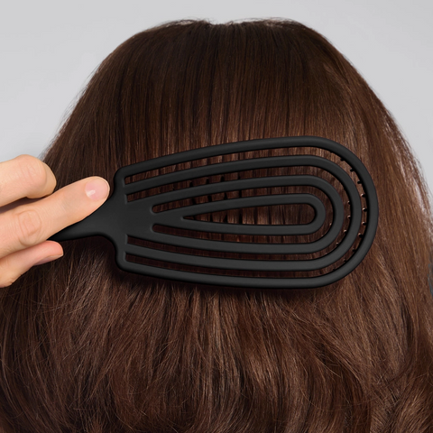 Person using a black hairbrush on their brown hair against a gray background