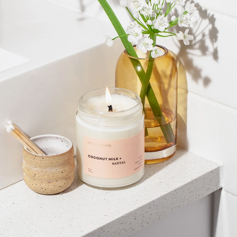 Candle with a pink label next to a vase with flowers on a white surface
