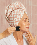 Woman with a checkered towel on her head using a black facial brush in a bathroom setting.