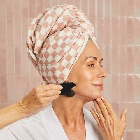 Woman with a checkered towel on her head using a black facial brush in a bathroom setting.
