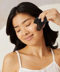 Woman using a black facial cleansing brush on her face with a neutral background